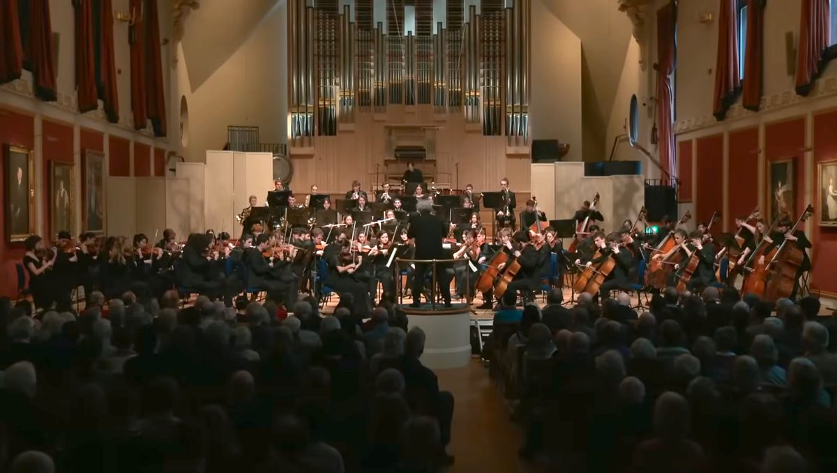 A conductor and symphony orchestra seen from the middle aisle of a busy audience.