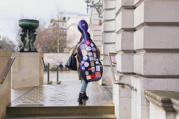 A student carrying a cello case covered in stickers walks into the Academy front entrance.