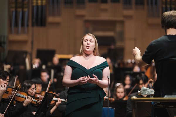 A solo vocal performer stands in front of an orchestra mid-performance.