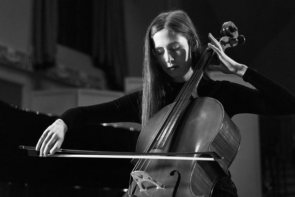 A student plays the cello alone with eyes closed in high contrast lighting (black and white).
