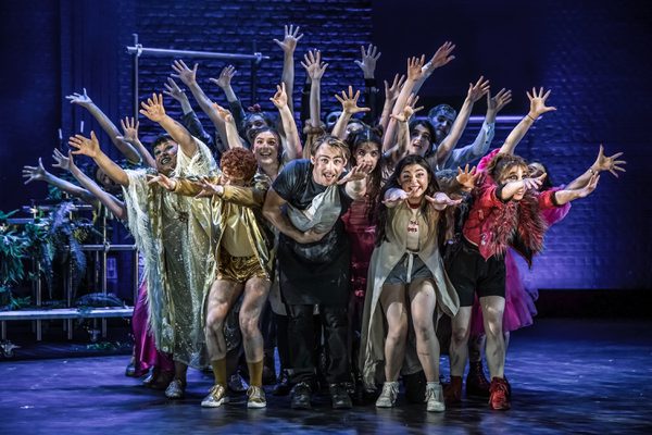 A cast of Musical Theatre performers on stage with hands raised high.