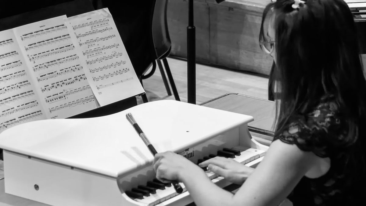 A toy piano player strikes the lid with a drumstick.