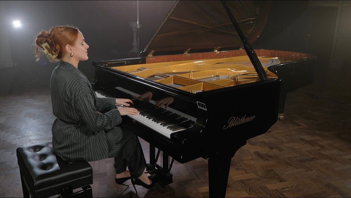 A student sits at a piano with the lid open, playing the keys against a heavy backlight.