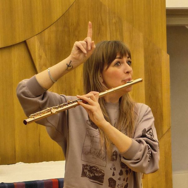Abigail Burrows holding a flute to mouth, motioning upwards with the other hand.