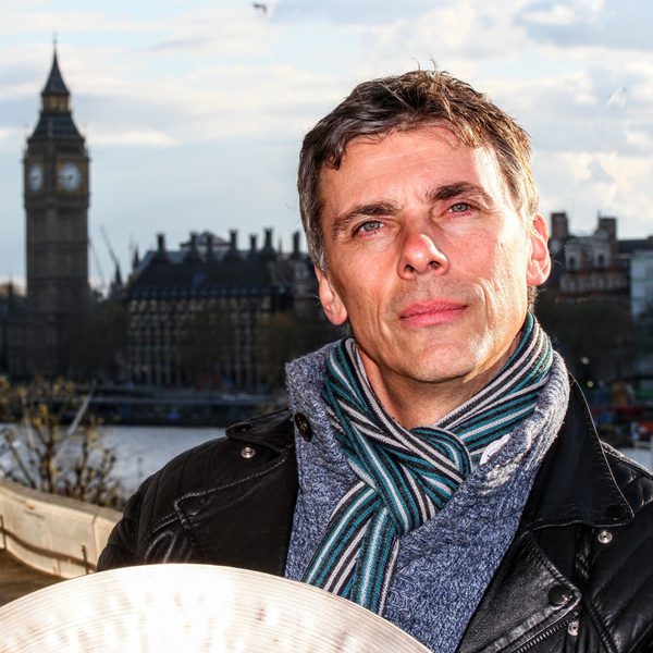 Andrew Barclay stands holding a cymbal with the Houses of Parliament in the background.