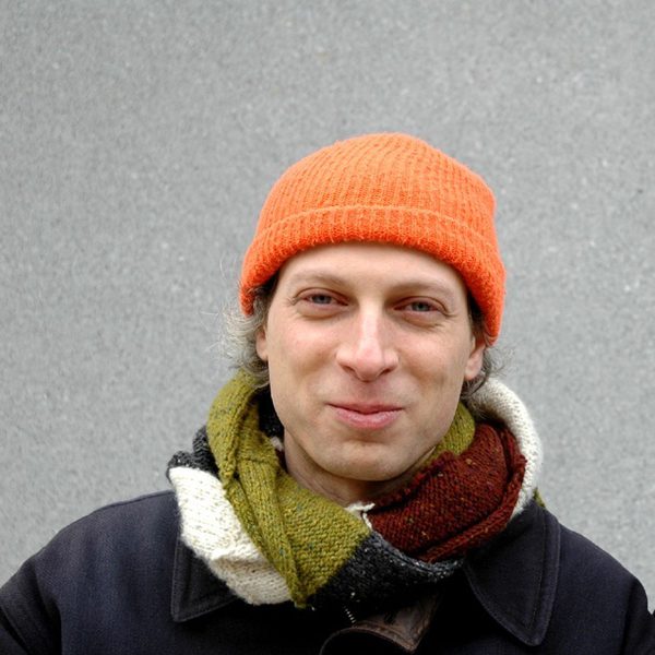 Barak Schmool stands against a grey background and smiles to camera, wearing an orange woolly hat and an autumnal scarf.