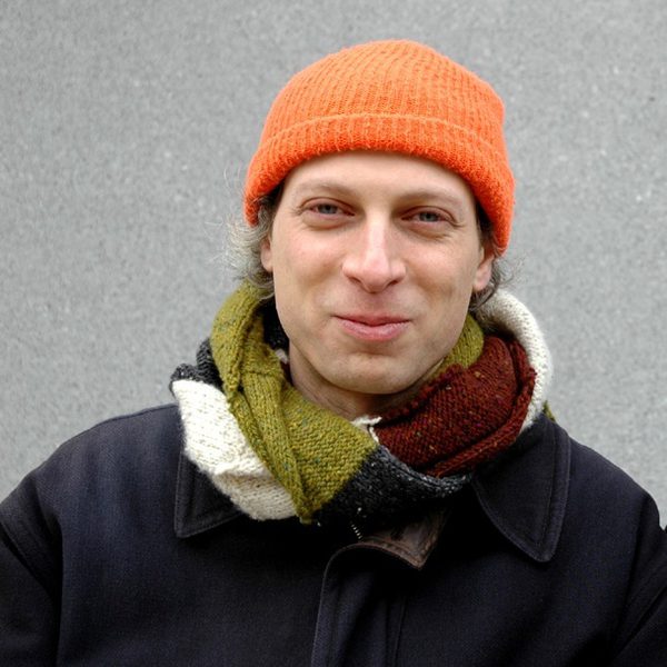 Barak Schmool stands against a grey background and smiles to camera, wearing an orange woolly hat and an autumnal scarf