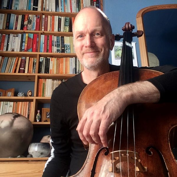 Ben Davies poses facing camera in an indoor study, holding cello.
