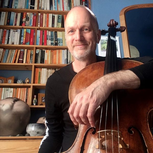 Ben Davies poses facing camera in an indoor study, holding cello