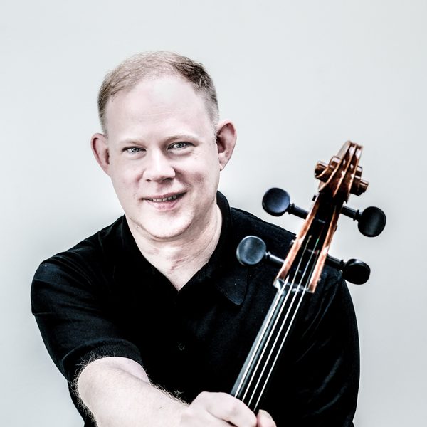 Portrait of Benjamin Hughes holding the neck of a cello, smiling for camera.