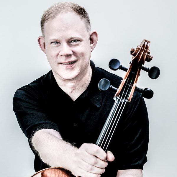 Portrait of Benjamin Hughes holding the neck of a cello, smiling for camera.