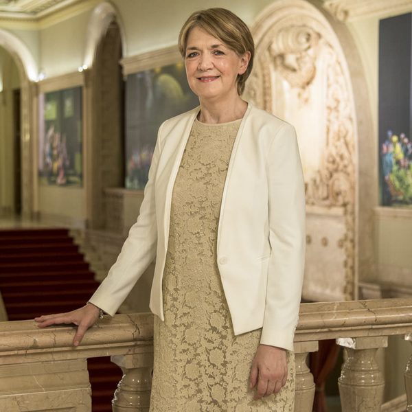 Brenda Hurley smiles to camera in a brightly-lit auditorium while holding a the balcony ledge.
