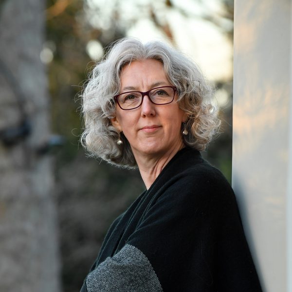 Cecelia Bruggemeyer stands with arms folded against a sunlit wall, wearing black and grey.
