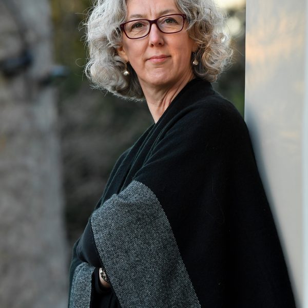 Cecelia Bruggemeyer stands with arms folded against a sunlit wall, wearing black and grey.