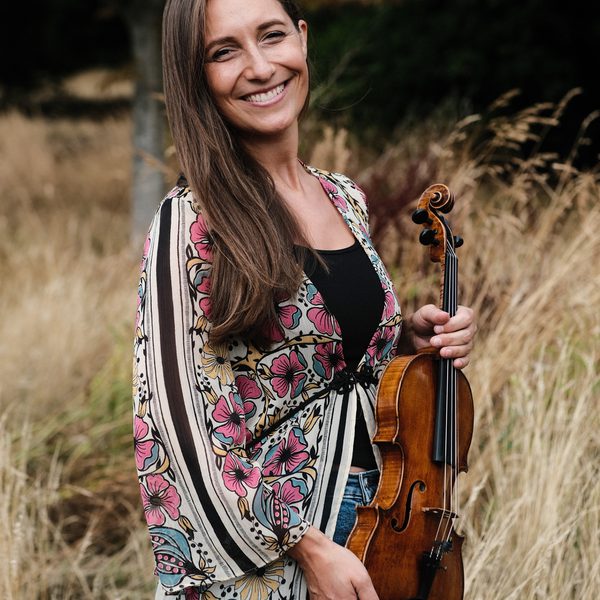 Portrait of Charis Jenson in a rural outdoor setting, holding a violin at waist height.