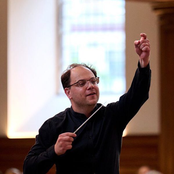 Long-distance portrait of Chris Hopkins conducting an orchestra, raising his non-baton hand.