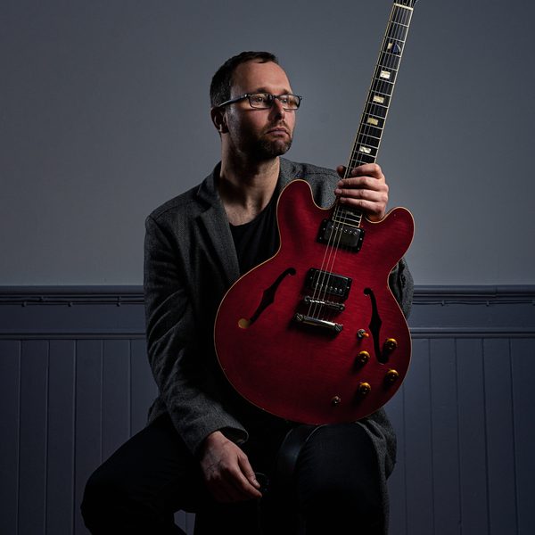 Portrait of Chris Montague sat on a stool, looking off camera and holding an electric guitar at the neck