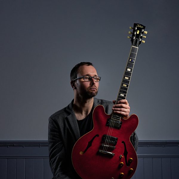 Portrait of Chris Montague sat on a stool, looking off camera and holding an electric guitar at the neck