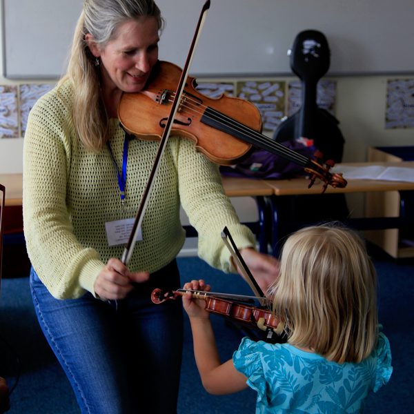 A smiling Karina MacAlpine stoops to teach a very young violin student