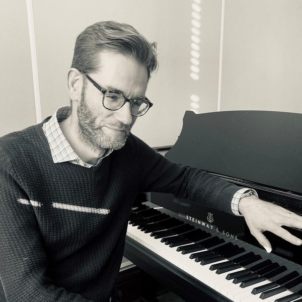 Black and white portrait of Daniel Swain Smiling, resting arm over the music stand on a piano keyboard.