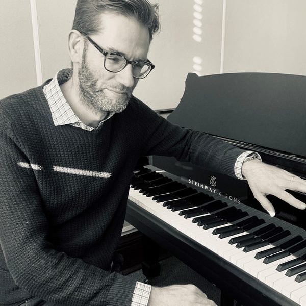 Black and white portrait of Daniel Swain Smiling, resting arm over the music stand on a piano keyboard