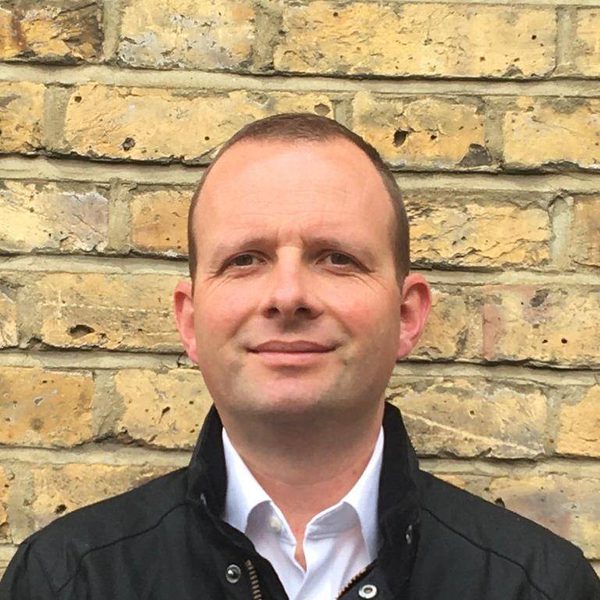 David Alexander stands posing for headshot, facing camera against a wall outside.