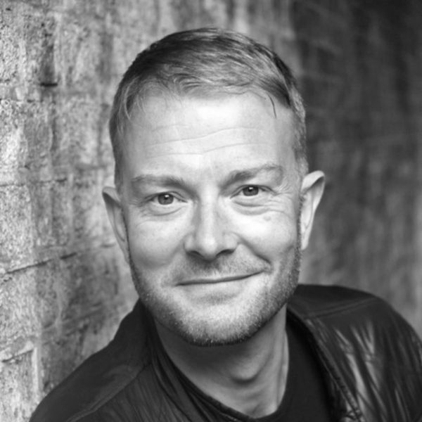 Portrait of David Ashley, smiling to camera while leaning against a brick wall.