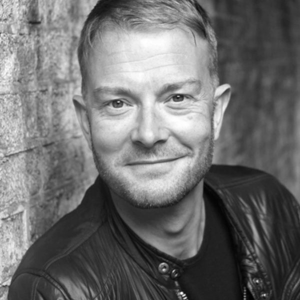 Portrait of David Ashley, smiling to camera while leaning against a brick wall