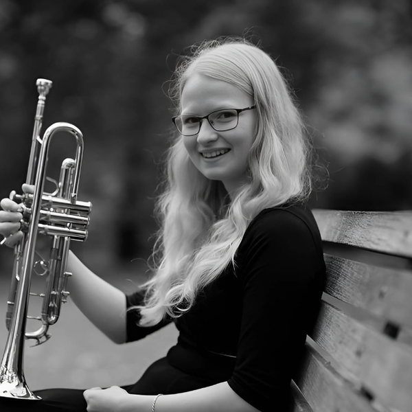 A black and white image of Emily Mitchell in profile, smiling to camera and sitting on a park bench holding a trumpet.