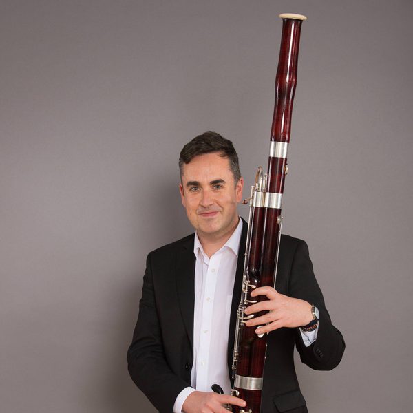 Wide portrait of Fraser Gordon smiling for camera in concert dress, holding a bassoon.
