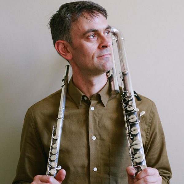 Gareth Lochrane holds two flutes, standing against a white wall