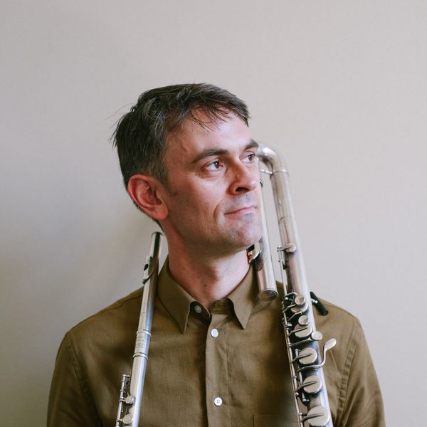 Gareth Lochrane holds two flutes, standing against a white wall.