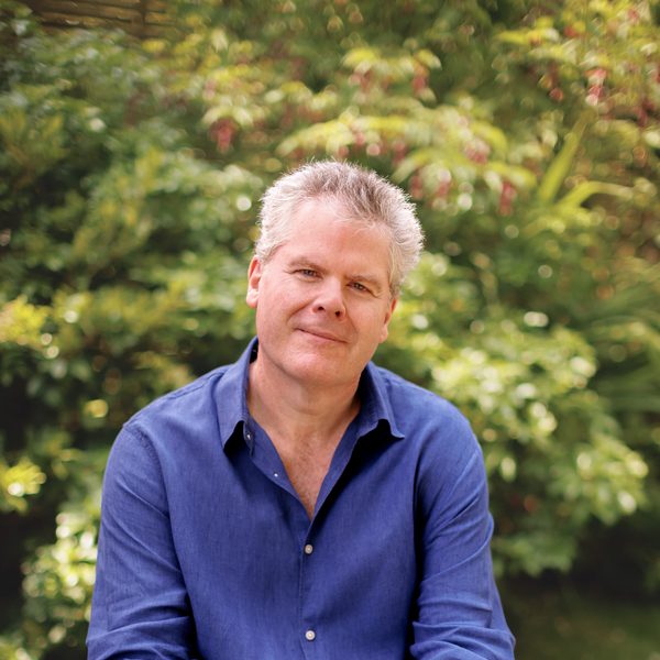 Gavin Carr sits on a bench looking to camera with hands clasped around one knee.