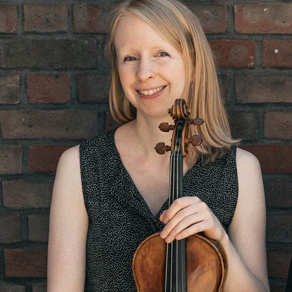 Gemma Sharples smiles for camera against a brick wall, holding a violin