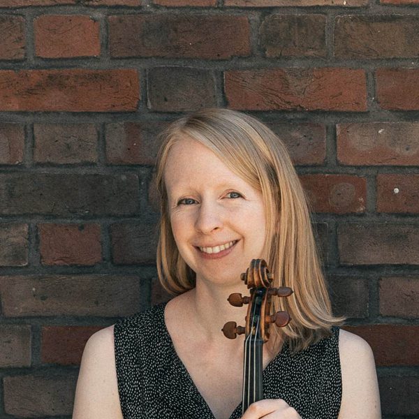 Gemma Sharples smiles for camera against a brick wall, holding a violin.