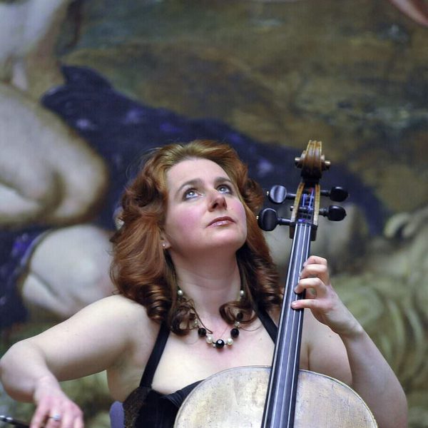Hannah Roberts plays cello in front of a large unspecified oil painting, gazing upwards.