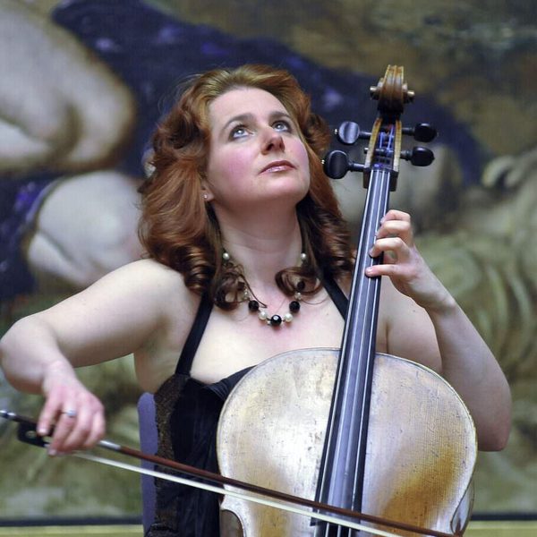 Hannah Roberts plays cello in front of a large unspecified oil painting, gazing upwards