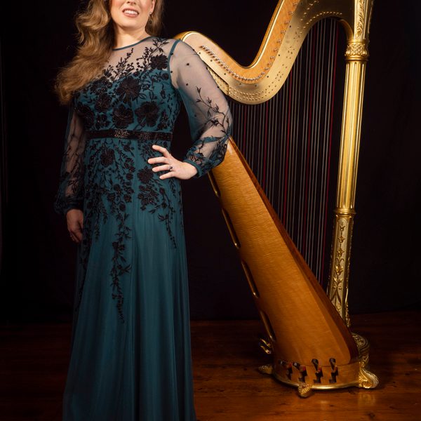 A harpist stands in front of a harp with left hand on hip