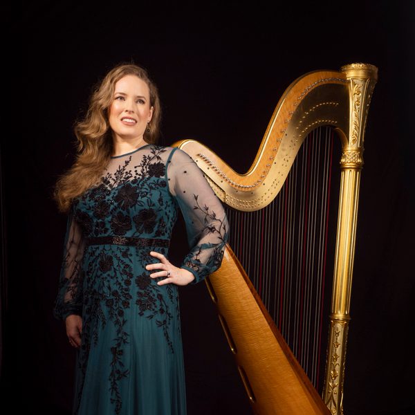 A harpist stands in front of a harp with left hand on hip.
