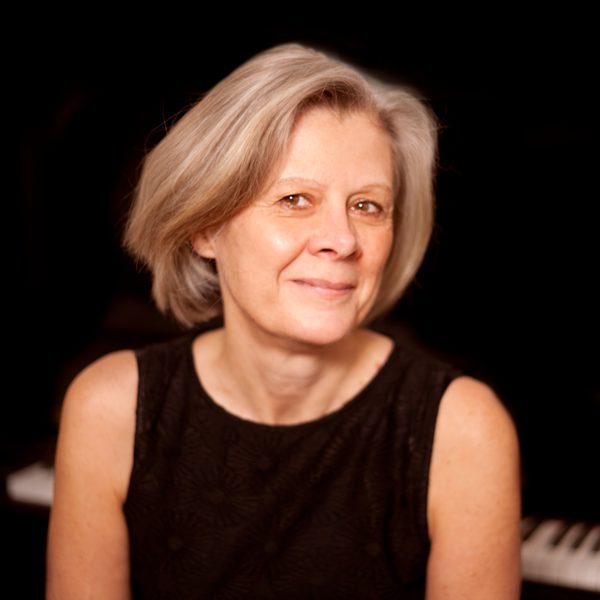 Helen Cawthorne sits at a piano, smiling for camera against a black background.