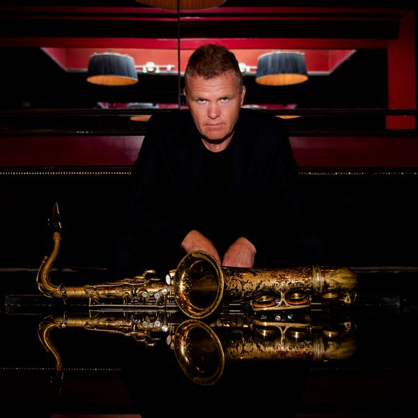 Portrait of Iain Bellamy looking to camera, with arms resting atop a saxophone placed on its side