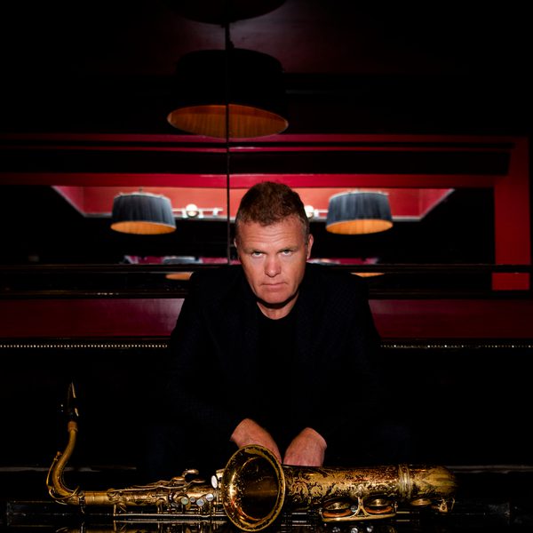 Portrait of Iain Bellamy looking to camera, with arms resting atop a saxophone placed on its side.