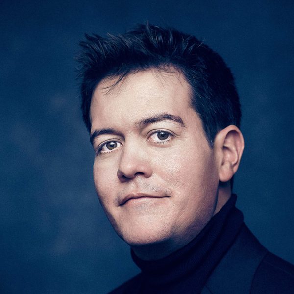 James Baillieu Smiles for headshot against a dark blue background.