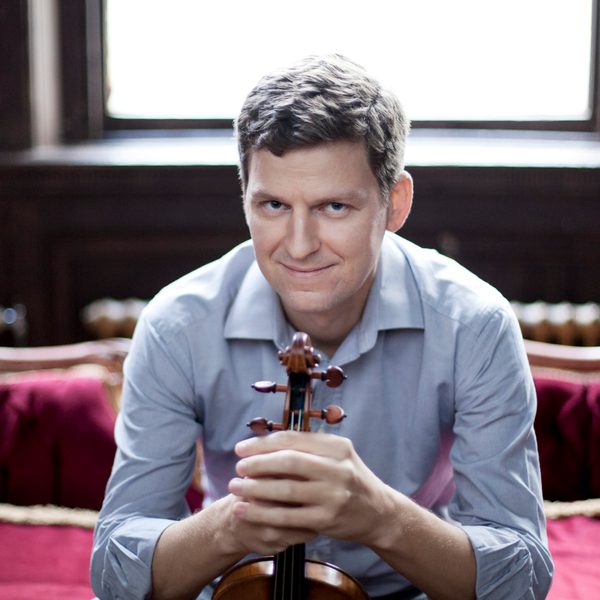 James Ehnes sits on a sofa, facing camera, smiling, holding a violin casually with both hands.