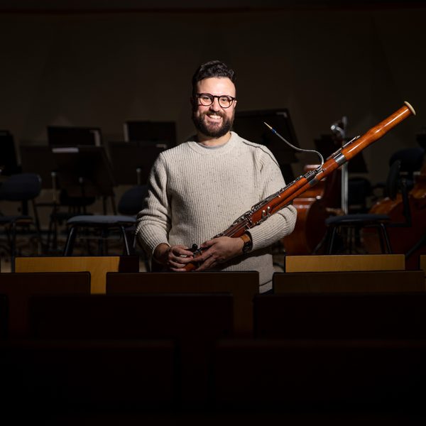 A man holds a bassoon, stood in a spotlight, smiling to camera.