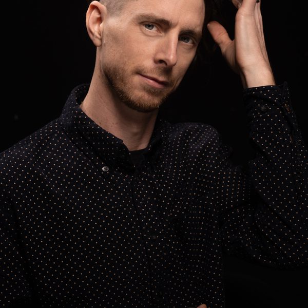 Portrait of Jim Hart looking to camera against a black background, running a hand through hair