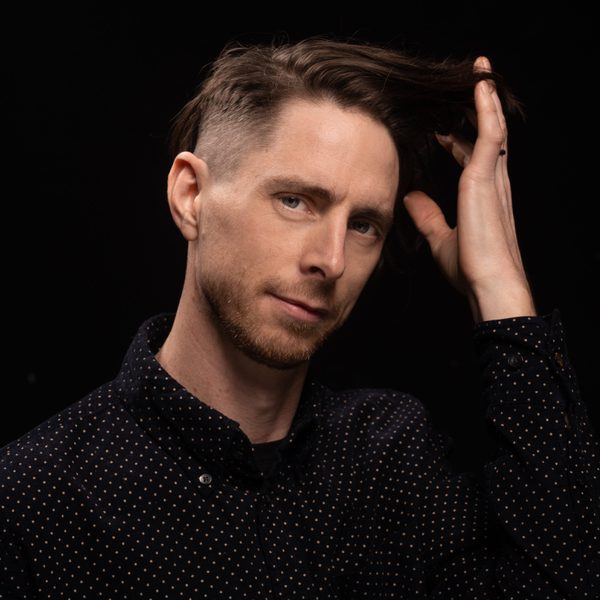 Jim Hart looking to camera against a black background, holding a hand against head.