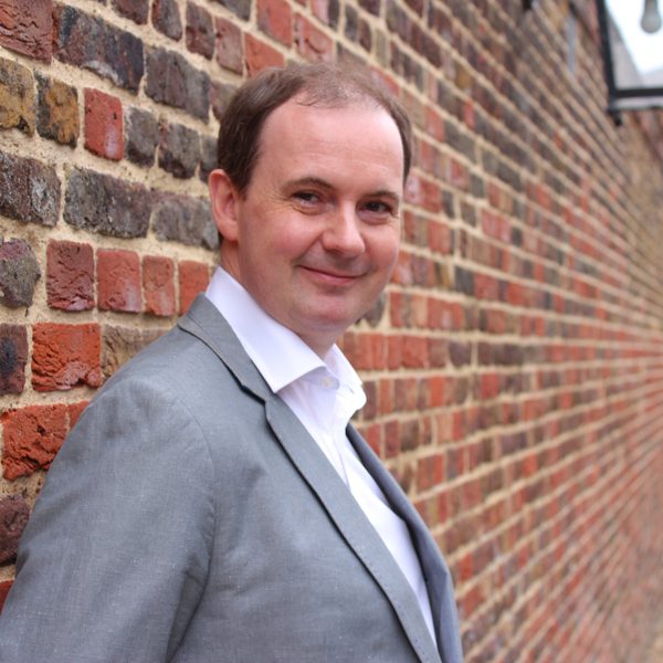 A smiling Joseph Tong stands in profile in a grey suit with his back to a brick wall.