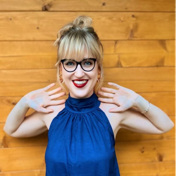 Julia Klimek stands against a wooden wall, smiling to camera in dark blue with elbows lifted.