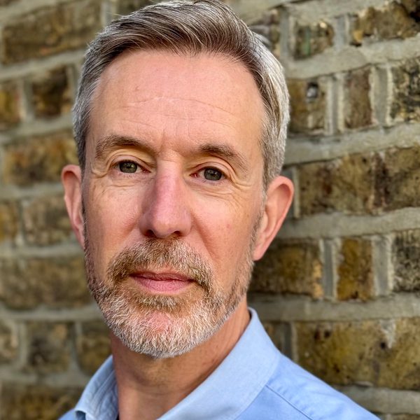 Portrait of Julian West looking to camera, smiling, against a brick wall.